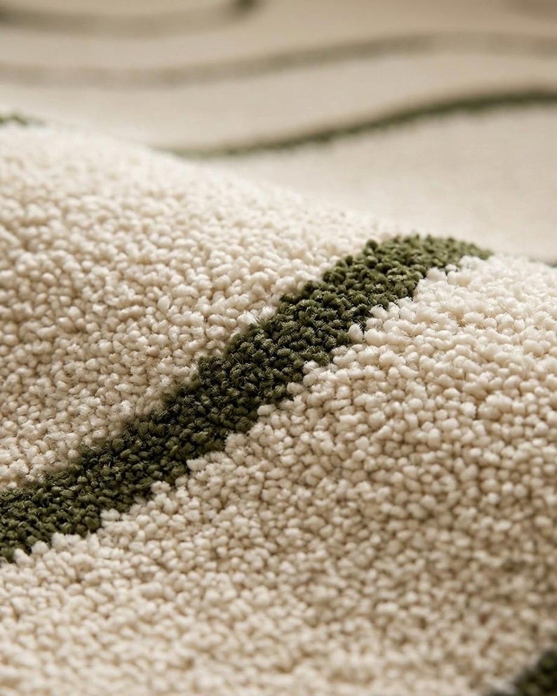 May include: A close-up view of a textured rug featuring a thick, cream-coloured pile with distinct, curved stripes in a deep olive green. The surface shows a dense, tufted construction with a soft, looped appearance throughout the fabric.