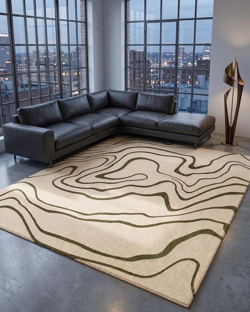 May include: A rectangular area rug featuring a cream-coloured base with dark olive green wavy, organic lines that mimic topographic map contours. The rug is positioned on a concrete floor in front of a large sectional sofa and a tall floor lamp.