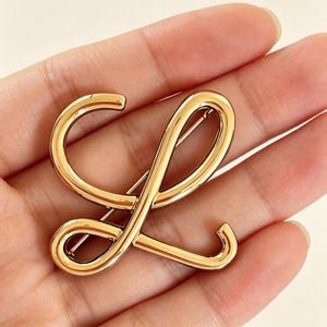 May include: A gold-colored brooch in the shape of the letter "L". The brooch has a smooth, polished finish and a pin on the back for attachment. The design is a cursive style, with rounded edges and a flowing form.
