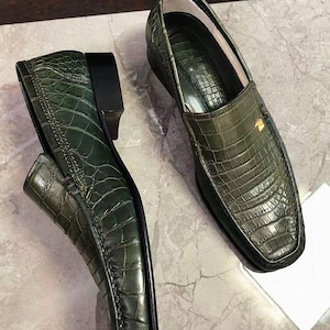 May include: A pair of dark olive green loafers featuring a textured crocodile skin pattern. Each shoe has a square toe, a low black heel, and a small gold-toned metal emblem attached to the side of the upper vamp near the opening of the shoe.