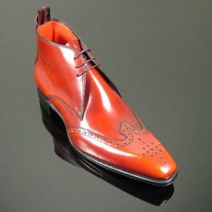 May include: A polished reddish-brown leather ankle boot featuring a pointed toe with decorative brogue detailing, a three-eyelet lace-up closure, a vibrant orange interior lining, and a black pull tab at the heel with visible white text.