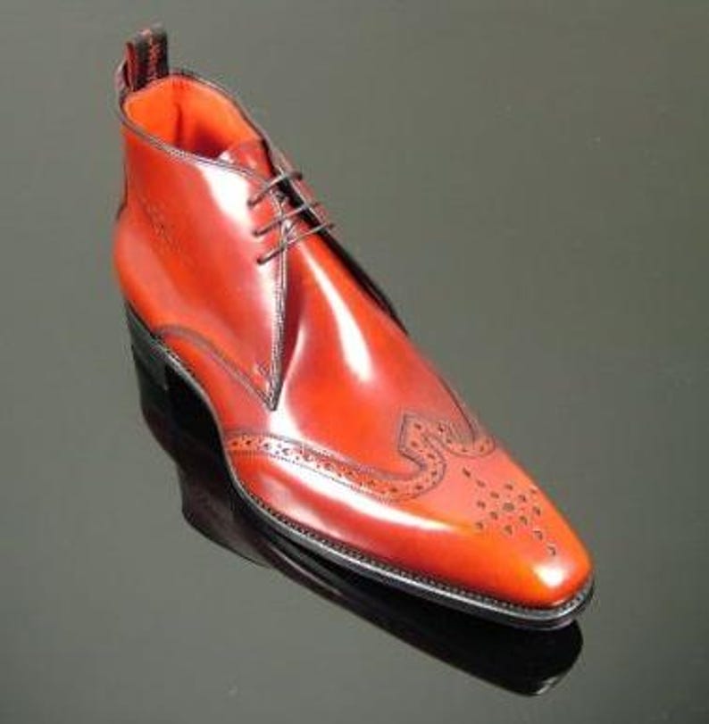 May include: A polished reddish-brown leather ankle boot featuring a pointed toe with decorative brogue detailing, a three-eyelet lace-up closure, a vibrant orange interior lining, and a black pull tab at the heel with visible white text.