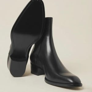 May include: Black leather ankle boots with a low block heel. One boot is angled to show the sole, revealing a stitched design. The other boot stands upright, showcasing the smooth, glossy finish of the leather. The boots have a classic, streamlined design.