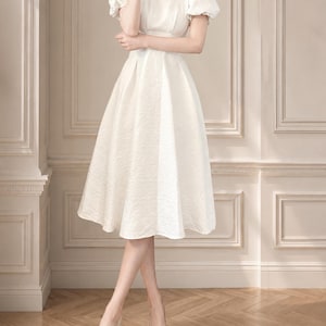May include: A white midi dress featuring a square neckline, short puff sleeves, and a textured fabric finish. The dress has a fitted bodice that transitions into a flared, pleated skirt that falls below the knees, paired with neutral-toned pointed heels.