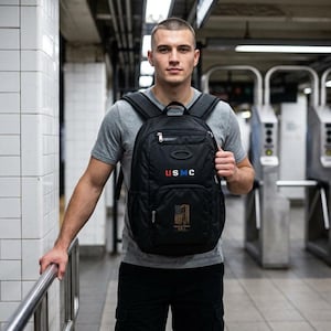 May include: Black backpack with the letters "USMC" in red, white, and blue. The backpack has multiple compartments and a design of the American flag. The backpack is being held by a person in a gray t-shirt.