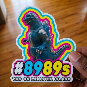 May include: A sticker featuring a blue Godzilla-like monster with a white, pink, yellow, and blue outline. The sticker has the text "#8989s VHS ON MONSTER ISLAND" in a bold, colorful font. The sticker is held by a hand.
