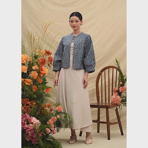 May include: A woman wearing a blue patterned jacket over a beige dress. The jacket has a geometric design. The dress is a solid color and reaches the ankles. The woman is standing in front of a floral arrangement and a wooden chair.