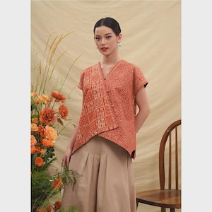 Op de afbeelding: Een roestkleurige wikkel top met een geometrisch patroon en korte mouwen. De top wordt gecombineerd met een beige broek met wijde pijpen. De outfit staat tegen een neutrale achtergrond met bloemstukken en een houten stoel.