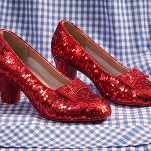 Puede incluir: Un par de zapatos de tacón de color rojo brillante cubiertos de lentejuelas, con punta redondeada, tacón de bloque y un lazo decorativo en el empeine. Los zapatos están recubiertos de lentejuelas rojas relucientes que reflejan la luz.