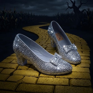 May include: A pair of silver pumps completely covered in reflective, sparkling rhinestones. Each shoe features a prominent, rectangular rhinestone-encrusted bow on the toe and a sturdy block heel. The interior of the shoe contains a small rectangular label.