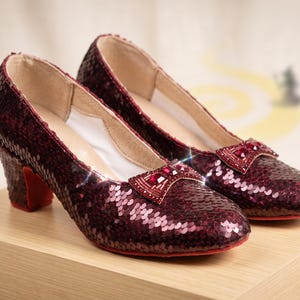 Puede incluir: Un par de zapatos de tacón de lentejuelas color burdeos oscuro con punta redondeada, tacón de bloque de altura media y un lazo decorativo en el empeine adornado con pequeños cristales rojos. El forro interior es blanco y las suelas son de color rojo vivo.