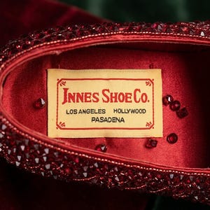 May include: Close-up of a red shoe interior with a label that reads "Innes Shoe Co." with locations in Los Angeles, Hollywood, and Pasadena. The shoe is embellished with red rhinestones.