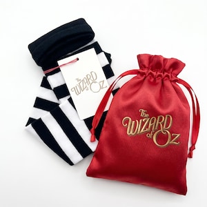 May include: Black and white striped socks with a white tag featuring the text The Wizard of Oz in gold, paired with a red satin drawstring pouch that displays the gold text The Wizard of Oz on the front, creating a themed set of accessories.