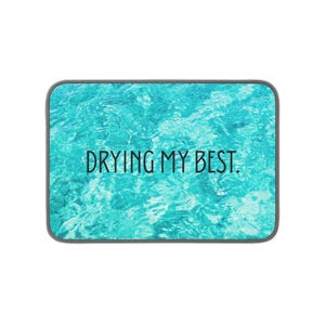 May include: A rectangular dish drying mat featuring a vibrant turquoise and light blue water ripple pattern. The text DRYING MY BEST. is printed in a simple black sans-serif font across the center of the mat, which has a dark gray border.
