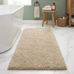 May include: A rectangular, beige bath mat with a plush, shaggy texture. The mat is placed on a white marble tile floor in a bathroom setting. The mat is near a white bathtub.