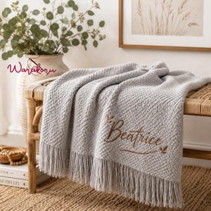 Può includere: Una coperta in maglia testurizzata grigio chiaro con lunghe frange sul bordo inferiore. La coperta è ricamata con il nome Beatrice in corsivo marrone, arricchito da piccoli disegni di foglie sopra e sotto il testo.