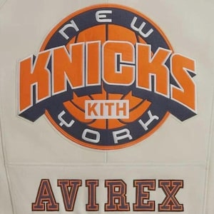 Puede incluir: Una chaqueta de color crema con un gran parche circular en la parte posterior. El parche muestra un gr&aacute;fico de baloncesto con el texto NEW YORK KNICKS y KITH en naranja y azul marino. Debajo del parche, la palabra AVIREX est&aacute; bordada en azul marino.