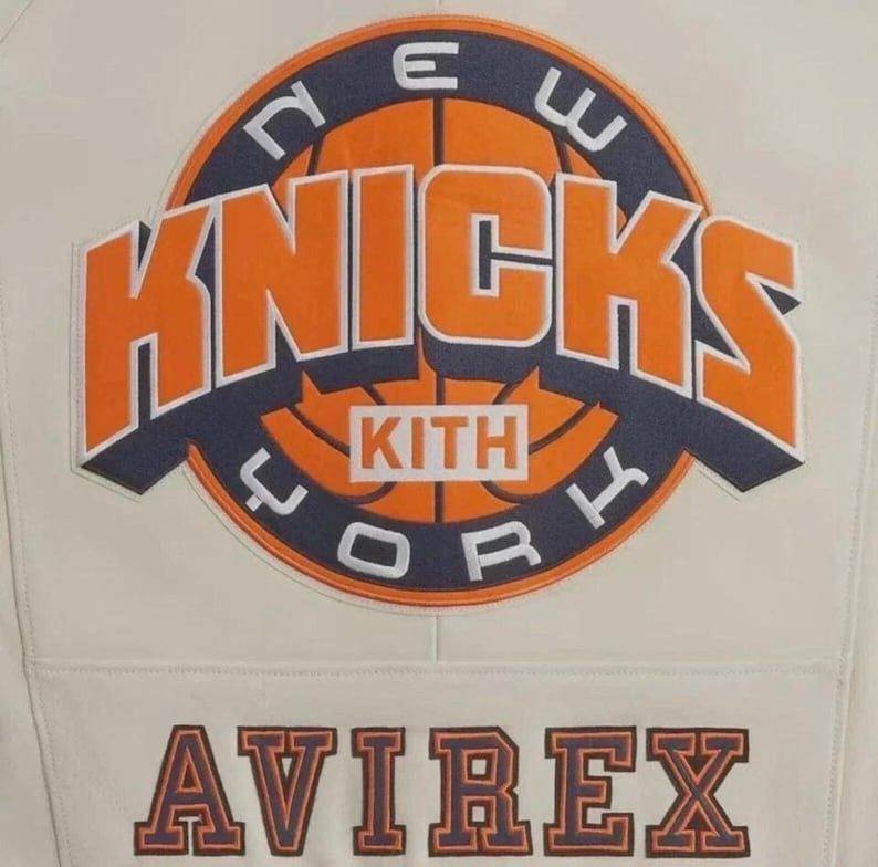 Puede incluir: Una chaqueta de color crema con un gran parche circular en la parte posterior. El parche muestra un gr&aacute;fico de baloncesto con el texto NEW YORK KNICKS y KITH en naranja y azul marino. Debajo del parche, la palabra AVIREX est&aacute; bordada en azul marino.