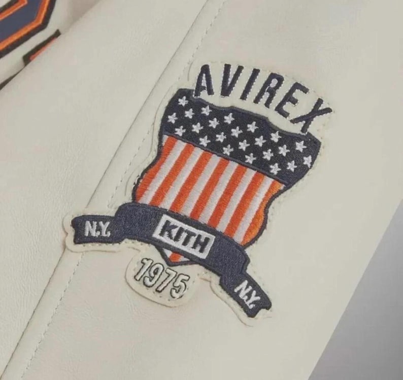 Puede incluir: Una chaqueta de cuero blanca con un parche bordado. El parche muestra el texto AVIREX, KITH, 1975 y N.Y. Incluye un gr&aacute;fico en forma de escudo con un campo azul de estrellas blancas sobre rayas verticales rojas y blancas, bordeado en azul marino.