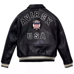 May include: A black leather jacket featuring the text AVIREX in white arched lettering across the upper back. Below this is a circular patch with a United States flag and the text ALL AMERICANS ESTABLISHED 1975. The word USA is printed below.