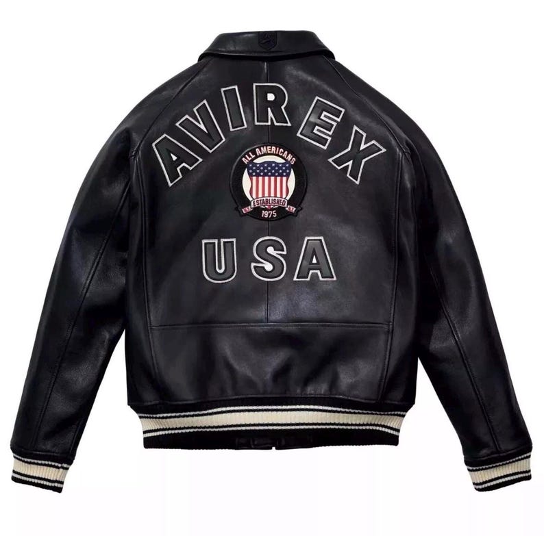 May include: A black leather jacket featuring the text AVIREX in white arched lettering across the upper back. Below this is a circular patch with a United States flag and the text ALL AMERICANS ESTABLISHED 1975. The word USA is printed below.