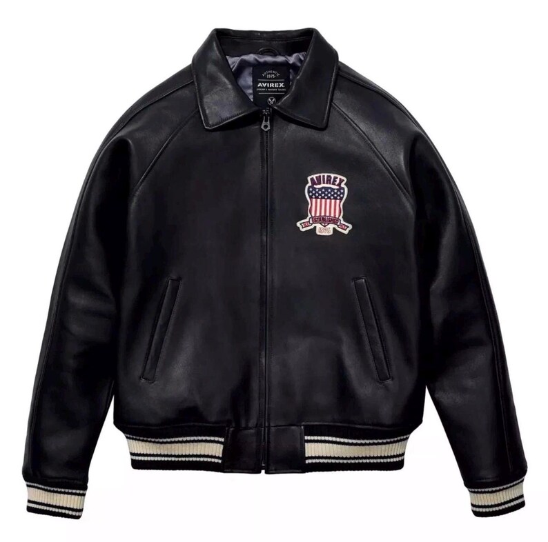 May include: A black leather jacket with a front zipper closure, a pointed collar, and two side pockets. The chest features an embroidered patch with the text AVIREX and a United States flag design. The cuffs and hem have black and white stripes.