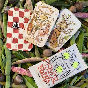 Puede incluir: Una vista cenital muestra tarjetas ilustradas y un pequeño paquete sobre un lecho de judías verdes frescas y nueces. Las tarjetas presentan dibujos caprichosos, incluido un diseño a cuadros rojos y blancos con la palabra "FRESH". El paquete dice "FLOWER FOLKS" y "3 CARD BOOSTER".