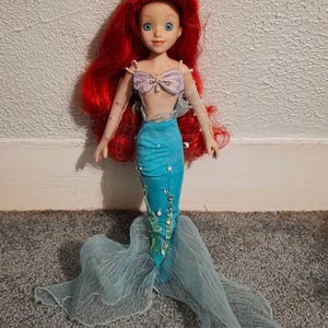 May include: A doll of a mermaid with long red hair, blue eyes, and a light complexion. She wears a lavender seashell top, a turquoise mermaid tail, and a flowing, sheer, light blue tail. The doll is standing on a gray carpet.