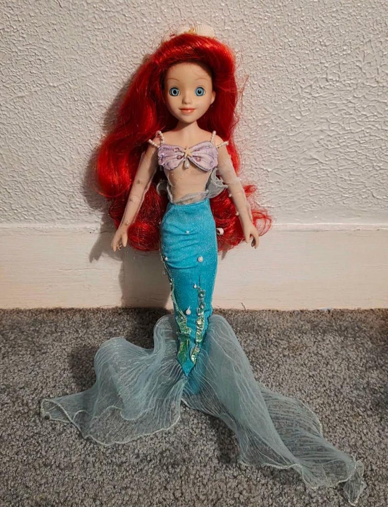 May include: A doll of a mermaid with long red hair, blue eyes, and a light complexion. She wears a lavender seashell top, a turquoise mermaid tail, and a flowing, sheer, light blue tail. The doll is standing on a gray carpet.