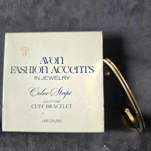 Può includere: Un bracciale a polsino color oro accanto alla sua scatola originale. La scatola bianca riporta la scritta "AVON FASHION ACCENTS IN JEWELRY" in nero. Il bracciale è di taglia media.