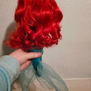 May include: A doll with vibrant red, curly hair and a teal tail. The doll's hair is styled in voluminous waves. The tail is made of a sheer, flowing fabric. The doll is held by a person wearing a light blue sweater.