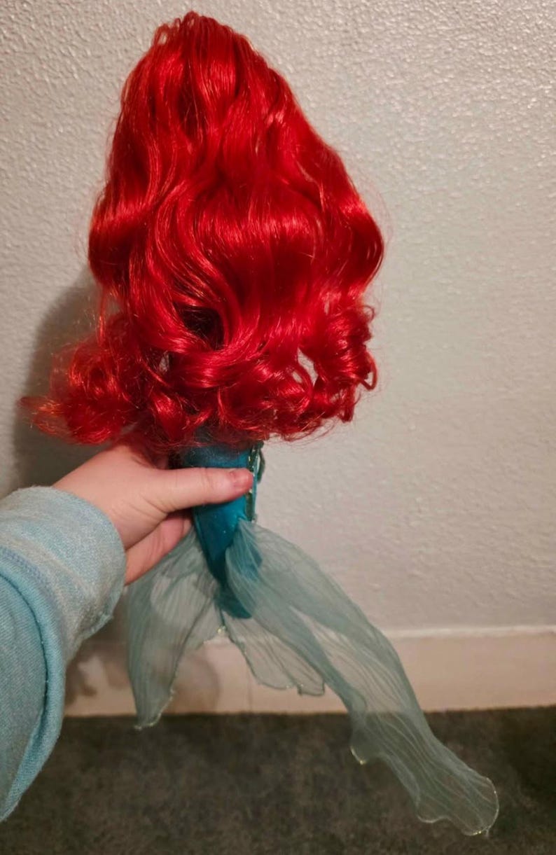 May include: A doll with vibrant red, curly hair and a teal tail. The doll's hair is styled in voluminous waves. The tail is made of a sheer, flowing fabric. The doll is held by a person wearing a light blue sweater.