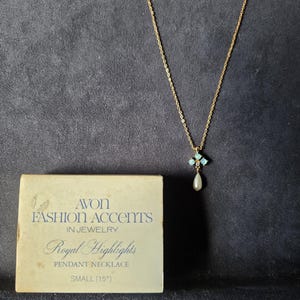 Könnte beinhalten: Eine goldfarbene Anhängerkette mit einem kleinen, quadratischen Anhänger mit türkisfarbenen Steinen und einer hängenden Perle. Die Halskette wird neben der Originalverpackung präsentiert, auf der steht: "AVON FASHION ACCENTS IN JEWELRY Royal Highlights PENDANT NECKLACE SMALL (15")", oder 38 cm.