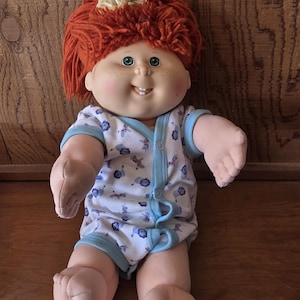 Peut inclure: Une poupée vintage avec des cheveux en fil rouge et des yeux verts. La poupée porte une combinaison blanche avec une bordure bleue et un motif de formes bleues et grises. La poupée a une expression souriante et est assise.
