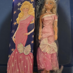 Pode incluir: Uma boneca de moda Jenna Avon de 29,2 cm num saco de plástico transparente ao lado da sua caixa azul original. A boneca tem cabelo loiro e veste um vestido rosa de um ombro só com um grande laço iridescente, luvas de renda e um colar.