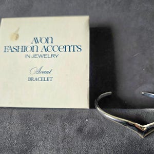 May include: A silver-toned Avant bracelet with a V-shaped design, next to its original box. The box is white with blue text that reads "AVON FASHION ACCENTS IN JEWELRY" and "Avant BRACELET".