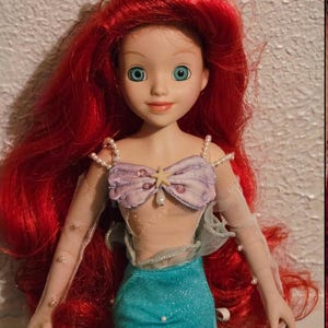 May include: A doll of Ariel, the Little Mermaid, with bright red hair and blue eyes. She wears a lavender seashell top, a turquoise skirt, and sheer sleeves adorned with pearls. A seashell crown sits atop her head.