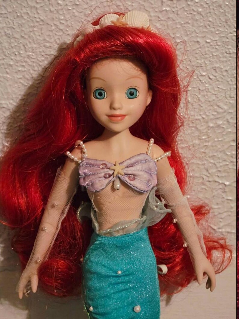 May include: A doll of Ariel, the Little Mermaid, with bright red hair and blue eyes. She wears a lavender seashell top, a turquoise skirt, and sheer sleeves adorned with pearls. A seashell crown sits atop her head.