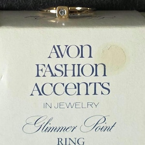 Op de afbeelding: Een goudkleurige ring met een kleine vierkante zetting met een enkele heldere steen, rustend op een witte doos bedrukt met de tekst AVON FASHION ACCENTS IN JEWELRY Glimmer Point RING in donkerblauwe letters met een beige sticker-afdruk.