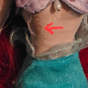 May include: A doll's bodice and skirt. The bodice is a light purple with a seashell design, adorned with beads and a starfish. The skirt is teal with a shimmering effect. Red hair is visible on the left side.