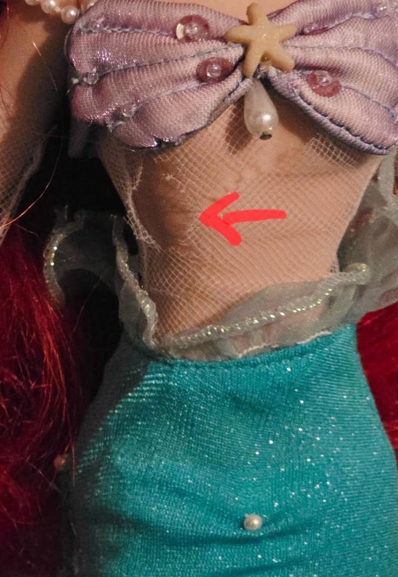 May include: A doll's bodice and skirt. The bodice is a light purple with a seashell design, adorned with beads and a starfish. The skirt is teal with a shimmering effect. Red hair is visible on the left side.