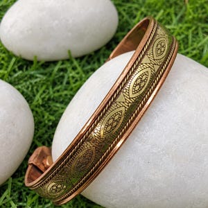 May include: A copper cuff bracelet featuring a central band of etched brass with a repeating geometric pattern of elongated ovals and triangles, bordered by two thin twisted metal ropes along the edges of the polished copper band.