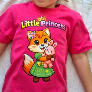 以下が含まれることがあります： 鮮やかなピンク色のTシャツで、黄色いリボンと髪に花をつけた緑色のドレスを着たキツネのキャラクターが描かれています。キツネは小さなピンク色のウサギを抱えています。キツネの上には、黄色とピンクの文字でLittle Princessとプリントされています。
