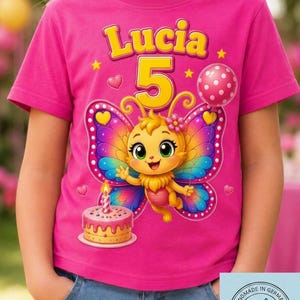 Op de afbeelding: Roze T-shirt met de naam "Lucia" en het nummer "5" in geel, met een cartoonvlinder met kleurrijke vleugels, een taart en een stippelballon. Het shirt is handgemaakt in Duitsland.