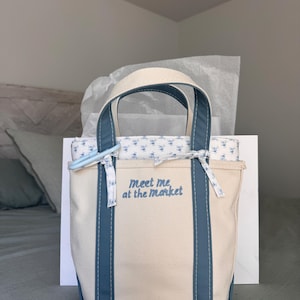 May include: A canvas tote bag with blue accents and handles. The bag has a cream-colored body with blue trim and the text "Meet me at the market" embroidered on the front. The bag is filled with a patterned fabric.