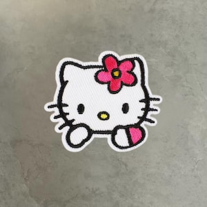 May include: An embroidered patch featuring the white face of a cartoon cat with black outlines, black oval eyes, a yellow oval nose, and six black whiskers. A bright pink flower with a yellow center is positioned above the left ear.