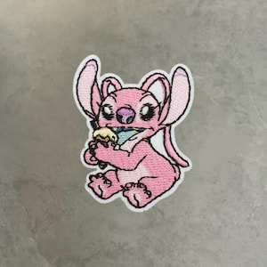 Peut inclure: Un écusson brodé représentant un personnage extraterrestre rose aux grandes oreilles, assis et tenant un cornet de glace multicolore. Le personnage a des yeux noirs, un nez violet et une bordure blanche soulignant toute la forme brodée.