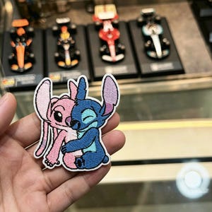 以下が含まれることがあります： 抱き合う2人のキャラクターが描かれた刺繍ワッペン。一方は長い耳を持つライトピンクのキャラクターで、もう一方は短い耳でウィンクをしているダークブルーのキャラクターです。ワッペンには白い縁取りと細かなステッチが施されています。