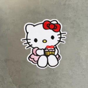 Puede incluir: Un parche bordado que muestra un personaje de gato blanco con un lazo rojo, bigotes negros y nariz amarilla. El personaje lleva un vestido rosa claro y sostiene un pequeño cupcake con glaseado rosa y base amarilla, delineado con hilo negro.