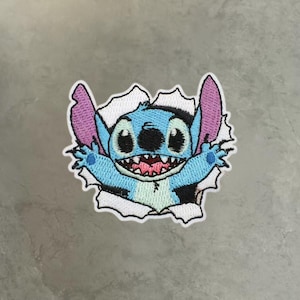May include: An embroidered patch featuring a light blue creature with large ears, black eyes, and a wide grin showing teeth, appearing to break through a white paper-like border with jagged edges, rendered in shades of blue, pink, and black.
